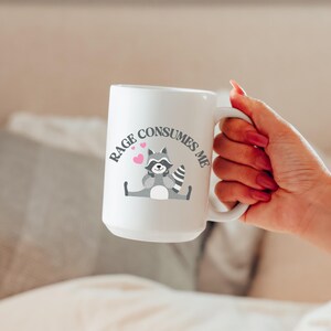 Peut inclure: Une tasse en c&eacute;ramique blanche avec une illustration de raton laveur noir et gris et le texte "RAGE CONSUMES ME" en lettres noires. Deux c&oelig;urs roses sont au-dessus du raton laveur.