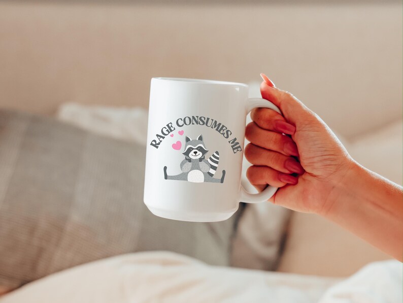 Peut inclure: Une tasse en c&eacute;ramique blanche avec une illustration de raton laveur noir et gris et le texte "RAGE CONSUMES ME" en lettres noires. Deux c&oelig;urs roses sont au-dessus du raton laveur.