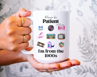 May include: White ceramic mug featuring retro icons like a floppy disk, cassette tape, and boombox. The mug has the text "Please be Patient with me, I'm from the 1900s" in a playful font, perfect for a nostalgic gift.