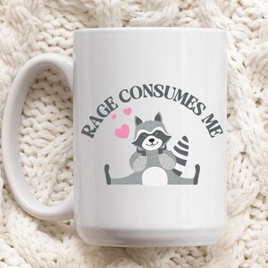 Peut inclure: Une tasse &agrave; caf&eacute; en c&eacute;ramique blanche avec une illustration de raton laveur noir et gris et le texte "RAGE CONSUMES ME".