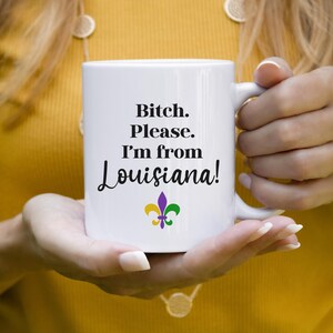 May include: White ceramic mug with the text "Bitch. Please. I'm from Louisiana!" in black script. The mug features a purple, gold, and green fleur-de-lis design. The mug is held by a person wearing a yellow shirt.