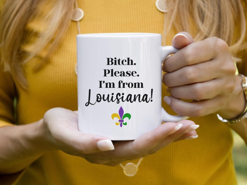 May include: White ceramic mug with the text "Bitch. Please. I'm from Louisiana!" in black script. The mug features a purple, gold, and green fleur-de-lis design. The mug is held by a person wearing a yellow shirt.