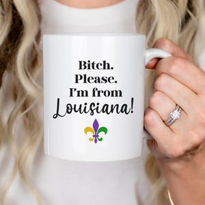 May include: White ceramic mug with the text "Bitch. Please. I'm from Louisiana!" in black, with a fleur-de-lis symbol in purple, yellow, and green. The mug is held by a person.