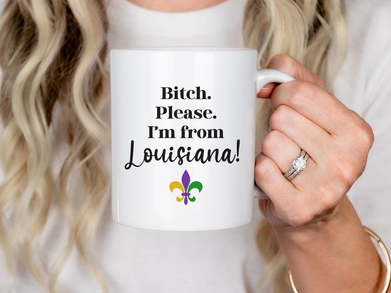 May include: White ceramic mug with the text "Bitch. Please. I'm from Louisiana!" in black, with a fleur-de-lis symbol in purple, yellow, and green. The mug is held by a person.