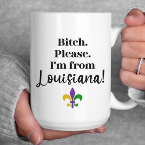 May include: White ceramic mug with the text "Bitch. Please. I'm from Louisiana!" in black script. A purple, gold, and green fleur-de-lis design is below the text. The mug is held by a person.