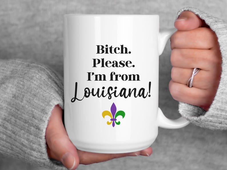 May include: White ceramic mug with the text "Bitch. Please. I'm from Louisiana!" in black script. A purple, gold, and green fleur-de-lis design is below the text. The mug is held by a person.