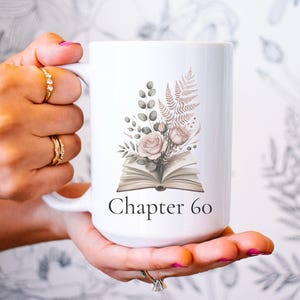 May include: White ceramic mug featuring a floral design with roses, greenery, and an open book. The mug has the text "Chapter 60" printed on it. The mug is held in a person's hand.