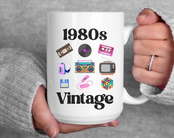 May include: White ceramic mug with the words "1980s Vintage" and illustrations of a cassette tape, vinyl record, boombox, and other retro items. The mug is held by a person wearing a gray sweater.