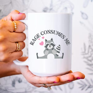 Peut inclure: Une tasse &agrave; caf&eacute; en c&eacute;ramique blanche avec une illustration de raton laveur gris et le texte "RAGE CONSUMES ME" en noir. Trois c&oelig;urs roses sont au-dessus du raton laveur.