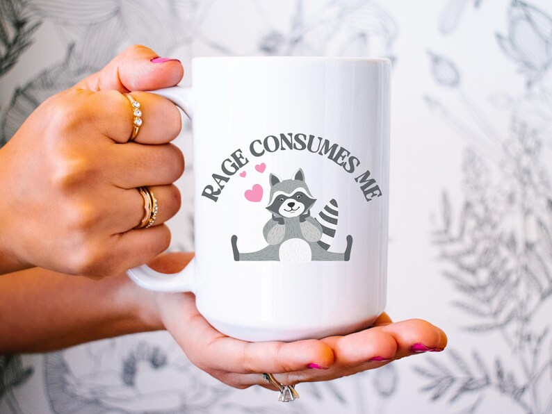 Peut inclure: Une tasse &agrave; caf&eacute; en c&eacute;ramique blanche avec une illustration de raton laveur gris et le texte "RAGE CONSUMES ME" en noir. Trois c&oelig;urs roses sont au-dessus du raton laveur.