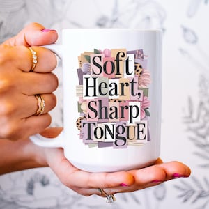 May include: White ceramic mug with the text "Soft Heart, Sharp Tongue" in a collage-style design with floral and leopard print accents. The mug is held by a person with gold rings.