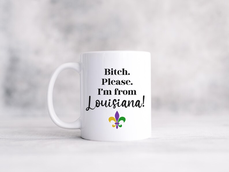 May include: White ceramic mug with the text "Bitch. Please. I'm from Louisiana!" in black script. A fleur-de-lis in purple, green, and gold is below the text. The mug has a white handle.