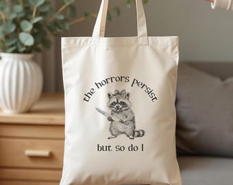 May include: A white canvas tote bag featuring a black and white illustration of a raccoon holding a knife. The text "the horrors persist but so do I" is printed on the bag.