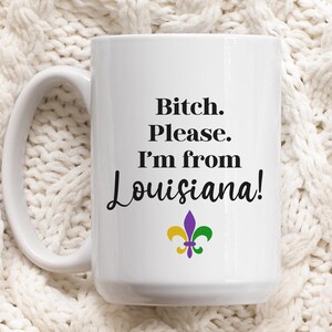 May include: White ceramic mug with the text "Bitch. Please. I'm from Louisiana!" in black script. A fleur-de-lis in purple, gold, and green is below the text. The mug has a large handle.