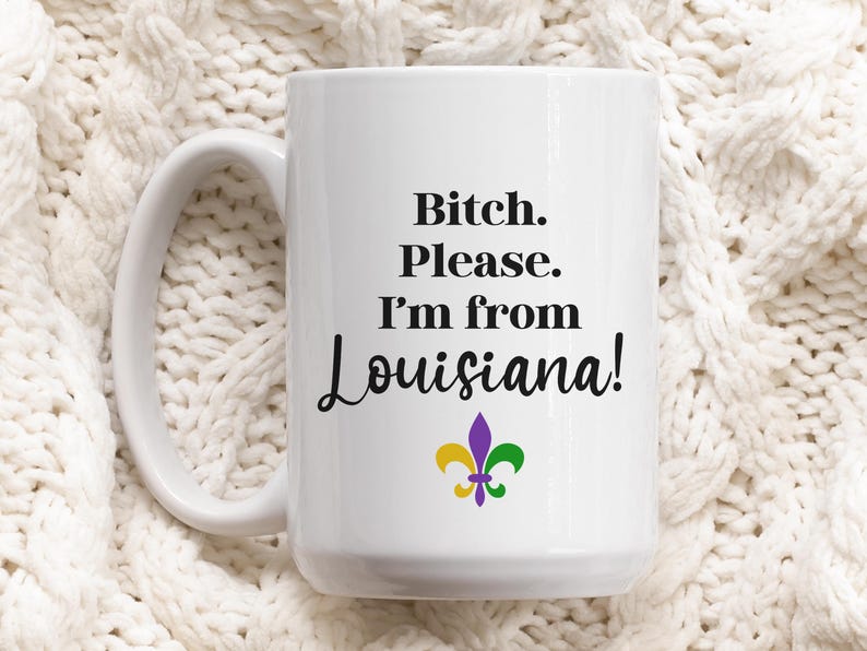 May include: White ceramic mug with the text "Bitch. Please. I'm from Louisiana!" in black script. A fleur-de-lis in purple, gold, and green is below the text. The mug has a large handle.