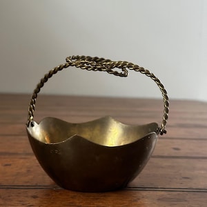 May include: A small, antique brass basket with a scalloped rim and a twisted handle. The basket is a warm, golden color and sits on a wooden surface. The handle is attached to the sides of the basket.