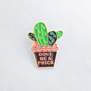 May include: A gold-colored enamel pin featuring a green cactus in a black pot with a red, white, and blue patterned rim. The text "Don't be a prick" is written in white on the black pot.
