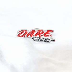 May include: A silver-colored pin with the word "D.A.R.E." in red lettering. Below the logo, the text "DRUGS ARE REALLY EXPENSIVE" is displayed in a smaller font. The pin is set against a white background.