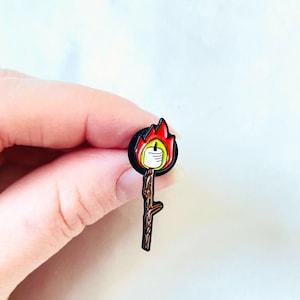 May include: A black enamel pin with a stick figure holding a lit candle with a red flame. The stick figure is wearing a white shirt and black pants.