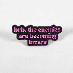 May include: Black enamel pin with pink text that reads "brb, the enemies are becoming lovers".