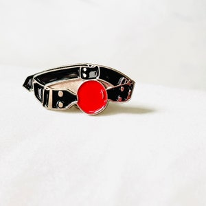 May include: A black and silver enamel pin featuring a red ball gag. The pin is a popular accessory for those who enjoy alternative fashion and accessories.