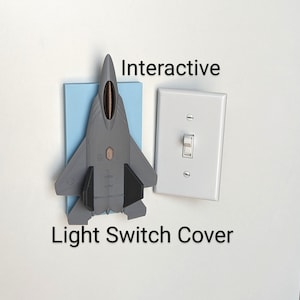 May include: A gray airplane-shaped light switch cover mounted on a light blue rectangle, with the text "Interactive Light Switch Cover". A white light switch is next to it.