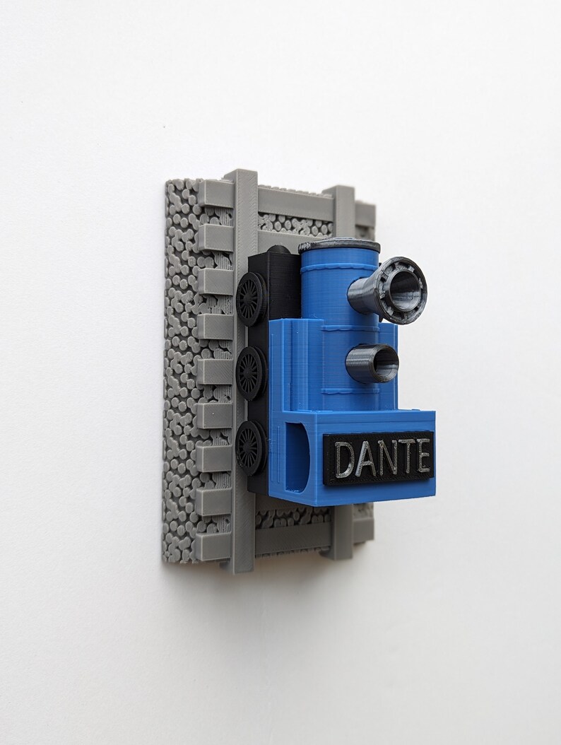 May include: A 3D-printed wall-mounted train ornament featuring a blue locomotive engine with a black nameplate reading DANTE. The train is positioned on grey plastic railroad tracks set against a textured grey base with a circular pattern.