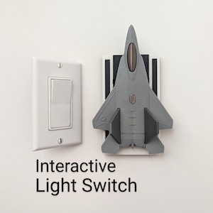 May include: A gray airplane-shaped light switch cover is mounted on a white wall next to a standard white light switch. The text "Interactive Light Switch" is below the switches.