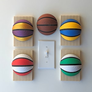 May include: Six colorful basketball-themed wall decorations. Each basketball is mounted on a beige square. The basketballs feature various color combinations: purple and yellow, brown, blue and yellow, red and white, and green and white.