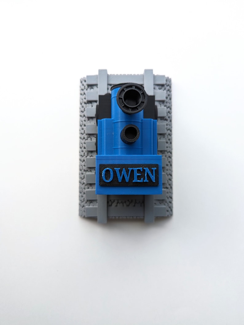 May include: A 3D printed blue and black toy train engine positioned on a gray textured railway track base. The front of the train features a black rectangular plate with the name OWEN embossed in blue capital letters, viewed from above.