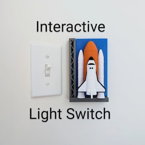 May include: White light switch next to a 3D-printed space shuttle design. The shuttle is white, orange, and blue, mounted on a gray base. The words "Interactive Light Switch" are printed above and below the objects.