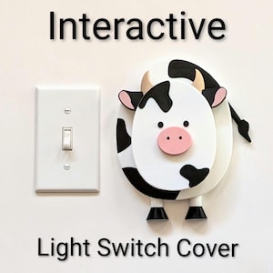 May include: A white light switch cover in the shape of a cow with black spots, a pink nose, and small horns. The words "Interactive Light Switch Cover" are printed above and below the cow. A white light switch is to the left.