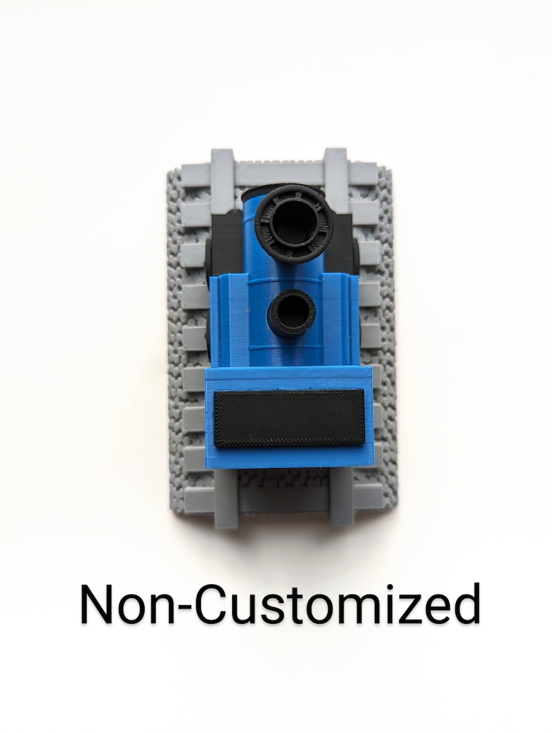 May include: A blue and black toy train engine positioned on a gray plastic track section with a textured base. The train features a prominent black chimney and a rectangular black plate on the front. The text Non-Customized is printed below.