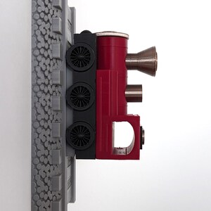 May include: A wall-mounted train locomotive model featuring a red body, black wheel assembly, and a grey textured track base. The engine includes a metallic funnel-shaped chimney and a cylindrical boiler section attached to a vertical grey rail.