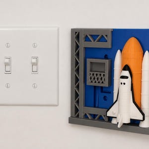 May include: A white double light switch plate next to a decorative wall plate featuring a 3D space shuttle launch scene. The scene includes a white and black shuttle, an orange fuel tank, white boosters, and a gray launch tower on a blue background.