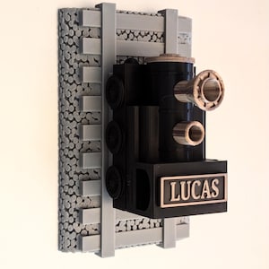 May include: A black 3D-printed train locomotive wall mount featuring the name LUCAS in raised lettering on the front. The train is attached to a gray textured base designed to resemble railroad tracks with gravel ballast underneath.