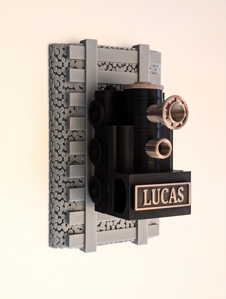 May include: A black 3D-printed train locomotive wall mount featuring the name LUCAS in raised lettering on the front. The train is attached to a gray textured base designed to resemble railroad tracks with gravel ballast underneath.