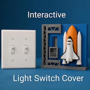 May include: A white double toggle light switch cover plate next to a decorative blue rectangular cover featuring a 3D space shuttle, orange fuel tank, and gray launch tower structure. The text Interactive Light Switch Cover is visible.