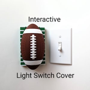 May include: A football-shaped light switch cover in brown and white, mounted on a green and white base, next to a standard white light switch. The image includes the text "Interactive Light Switch Cover".