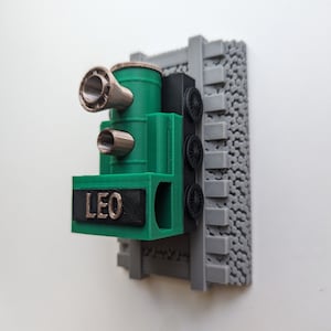 May include: A 3D printed green and black train engine wall mount featuring the name LEO in metallic lettering on the front. The train is positioned on a grey textured track base with three black wheels visible on the side and two metallic funnels.
