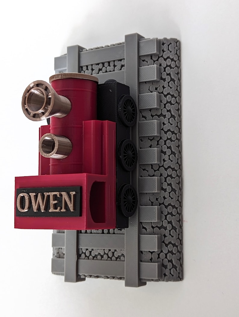 May include: A 3D printed train engine model in red, black, and metallic bronze, featuring the name OWEN in raised bronze letters on the front. The train is mounted on a gray plastic base designed to look like railroad tracks and gravel ballast.
