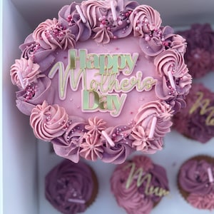 Può includere: Una torta glassata rosa e viola con le parole "Happy Mother's Day" in lettere dorate. La torta è circondata da cupcake con glassa e decorazioni simili.