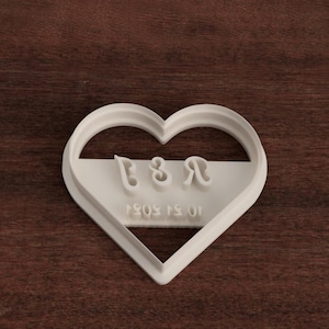 May include: White heart-shaped cookie cutter with the letters "B & R" and the date "15.01.2023" inscribed on it.