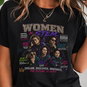 Pode incluir: Uma t-shirt preta com uma impressão gráfica intitulada WOMEN IN STEM. O design inclui retratos de Marie Curie, Katherine Johnson, Jane Goodall, Rosalind Franklin e Chien-Shiung Wu, com texto incluindo SMARTER. STRONGER. CHANGING. THE WORLD. e DREAM. DISCOVER. INNOVATE. THE FUTURE IS OURS.