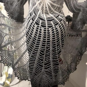 May include: A handmade, open-weave crochet cardigan in a spiderweb design. The garment features a gradient of white to gray, with flared sleeves and a long, flowing silhouette. The intricate pattern is visible from the back.