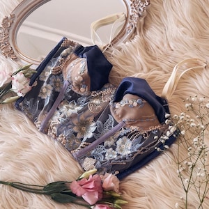 May include: A navy blue and beige embroidered corset with floral detailing and pearl accents. The corset features a sweetheart neckline and is adorned with delicate blue and beige flowers. The image also includes pink flowers and a decorative mirror.