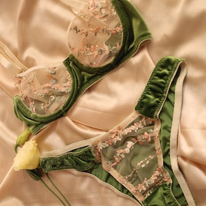 May include: A green velvet lingerie set, including a bra and matching briefs. The bra and briefs feature sheer panels with pink and green floral embroidery. The set is displayed on a satin surface.