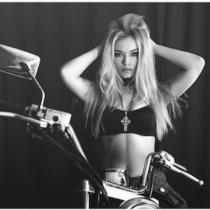 Puede incluir: Una foto en blanco y negro de una mujer con el pelo largo y rubio sentada en una motocicleta. Lleva una camiseta negra con un collar de plata con una cruz y una gargantilla negra.