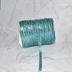 K&ouml;nnte beinhalten: Eine Spule mit tealblauem Glitzerband mit einem wei&szlig;en Etikett. Das Band ist um eine wei&szlig;e Pappspule gewickelt.