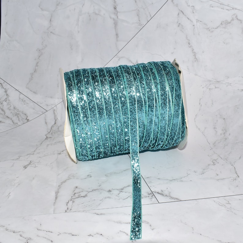 K&ouml;nnte beinhalten: Eine Spule mit tealblauem Glitzerband mit einem wei&szlig;en Etikett. Das Band ist um eine wei&szlig;e Pappspule gewickelt.
